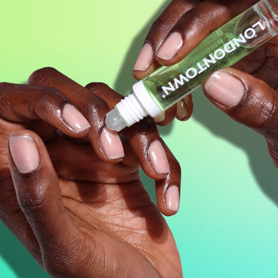 Hands applying the Roll & Glow Cuticle Oil - Agave Pear from a pocket-sized roll-on bottle labeled "LONDONTOWN" to manicured nails, against a gradient green background.