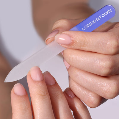 A close-up of two hands, one holding a lilac-handled glass nail file labeled "LONDONTOWN," while the other hand is being filed effortlessly. The nails are polished in light pink, ensuring nail filing without damage.