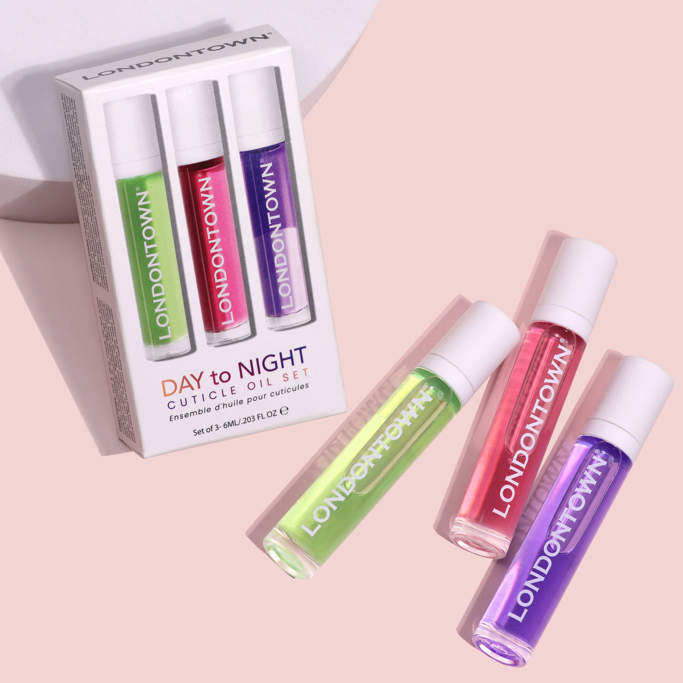LONDONTOWN's Day to Night Cuticle Oil Set showcases three vibrant cuticle oil bottles in green, purple, and pink, elegantly arranged partially outside their packaging on a pink background. This set is the perfect solution for dry or peeling cuticles.