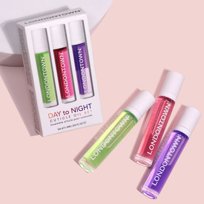 LONDONTOWN's Day to Night Cuticle Oil Set showcases three vibrant cuticle oil bottles in green, purple, and pink, elegantly arranged partially outside their packaging on a pink background. This set is the perfect solution for dry or peeling cuticles.