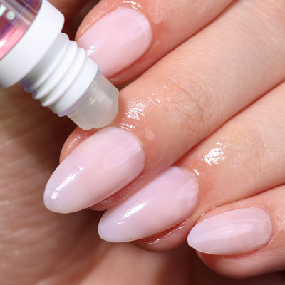 A close-up of a hand with pale pink manicured nails. A dropper is applying LONDONTOWN's Roll & Glow Cuticle Oil - Dragonfruit to the nails and cuticles, perfect for on-the-go care.