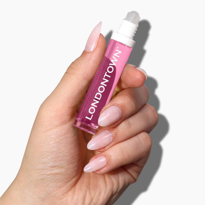 Close-up of a hand holding a pink LONDONTOWN Roll & Glow Cuticle Oil - Dragonfruit bottle against a white background. The hand, with long, pink, manicured nails, showcases this juicy cuticle hydrator perfect for on-the-go use.