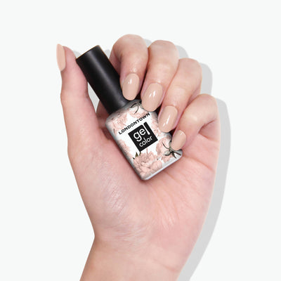 A hand with a nude manicure holds a bottle of LONDONTOWN Dusk gel color nail polish, showcasing an easy-to-wear, neutral peach shade.