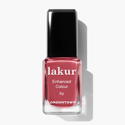 A bottle of LONDONTOWN Lakur nail polish in the shade Flushed Cheeks, showcasing a sun-kissed pinkish-red color and adorned with a black cap and white text on the front.