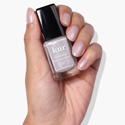 Hand holding a bottle of LONDONTOWN Quartz Illuminating Nail Concealer, the instant nail perfector. The nails are painted in a soft gray glaze.