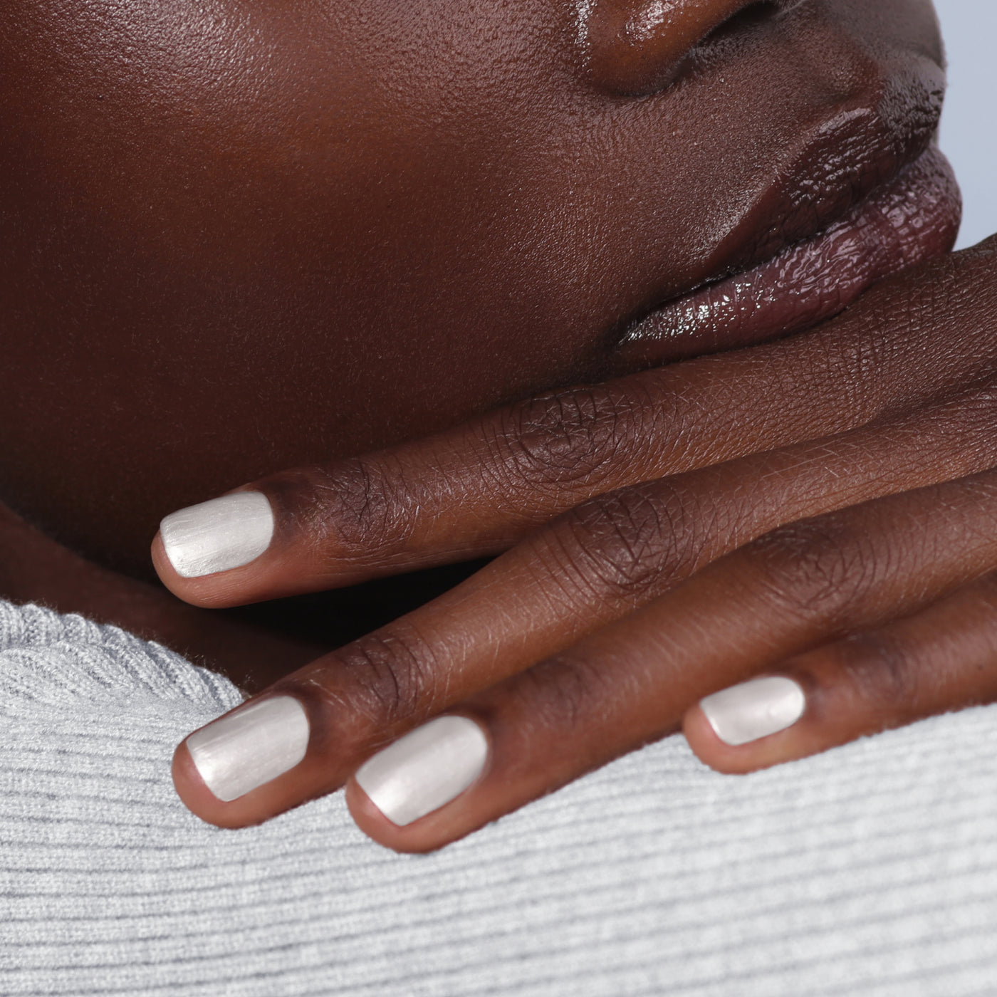 Close-up of a person's hand touching their face, with fingers showcasing LONDONTOWN's "Powder" nail polish—pearly white and reminiscent of powdered snow—resting against their lips. The person is adorned in a textured, light grey garment.