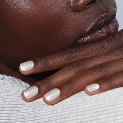 Close-up of a person's hand touching their face, with fingers showcasing LONDONTOWN's "Powder" nail polish—pearly white and reminiscent of powdered snow—resting against their lips. The person is adorned in a textured, light grey garment.