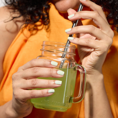 A person sporting an orange top holds a mason jar filled with Lichen green beverage from LONDONTOWN, sipping through a metal straw as the scent of white sage wafts gently in the background.