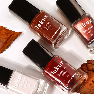 Four bottles of LONDONTOWN Lakur nail polish in various shades, including "Mull It Over," a decadent burgundy, and a metallic shade, are arranged on a white surface with scattered autumn leaves.