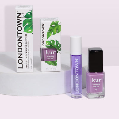 Image of LONDONTOWN's Lavender Dreams Duo nail care products, including Nighttime Cuticle Quench and two shades of Perfecting Nail Veil in bottles, all displayed on a white circular podium. The packaging features green leaf designs, adding a touch of natural perfection to your pampered routine.