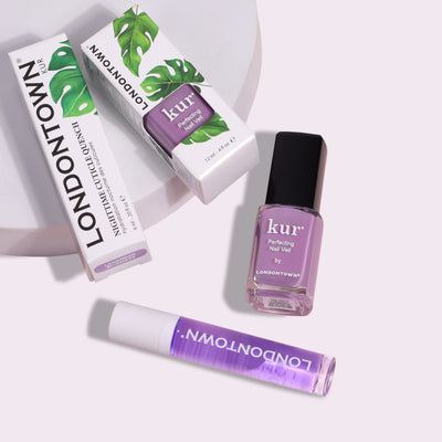 LONDONTOWN's Lavender Dreams Duo, featuring cuticle oil and nail polish, rest on a white surface. The cuticle oil's box showcases green leaf graphics, while the nail products display a pampered purple hue, ensuring your nails achieve perfection.