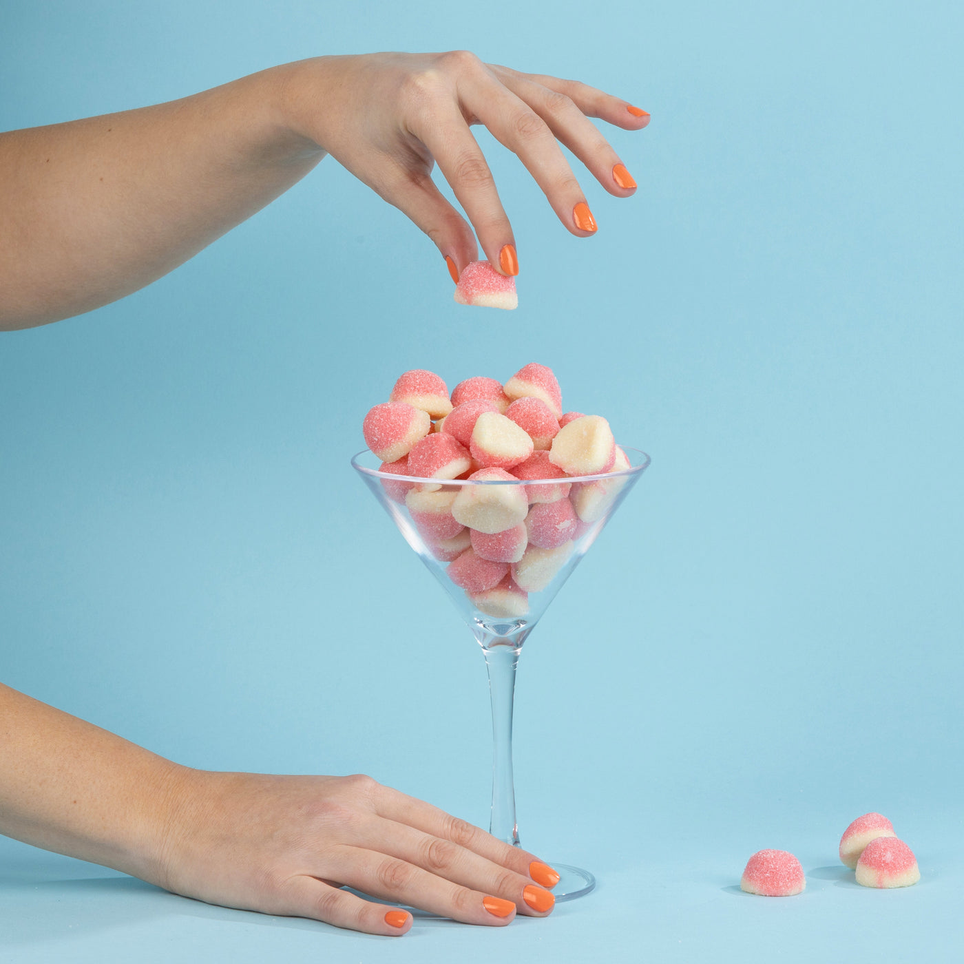 A hand with vibrant Paloma (Professional) orange-painted nails from LONDONTOWN holds a pink-and-white candy above a martini glass filled with similar candies, set against a blue background.