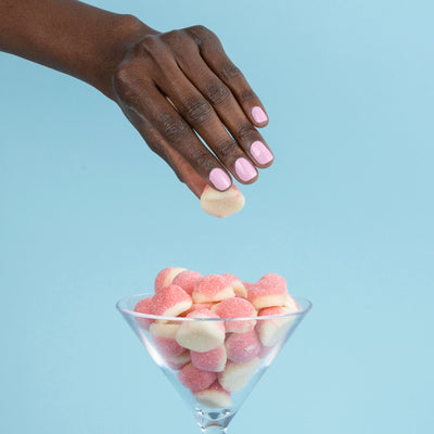 A hand adorned with LONDONTOWN's Pink Palm nails hovers over a glass filled with pink and white gummy candies, set against a blue background.