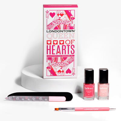 The LONDONTOWN Queen of Hearts Galentines Day Set features a nail file, brush, and two nail polishes in pink and peach, elegantly displayed on a white podium.