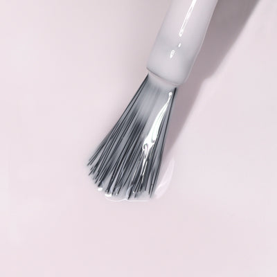 Close-up of the Ski Bunny nail polish brush from LONDONTOWN, with gray bristles applying clear, pink-tinted cream polish on a light surface.