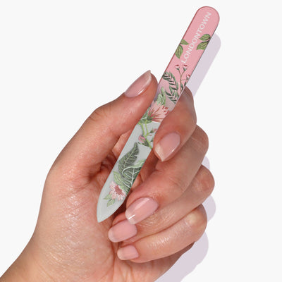 A hand holding an effortlessly chic, floral-patterned LONDONTOWN Glass Nail File - Safara with pink and white text on the handle. The nails are polished with a natural pink shade, ensuring a damage-free nail filing experience.
