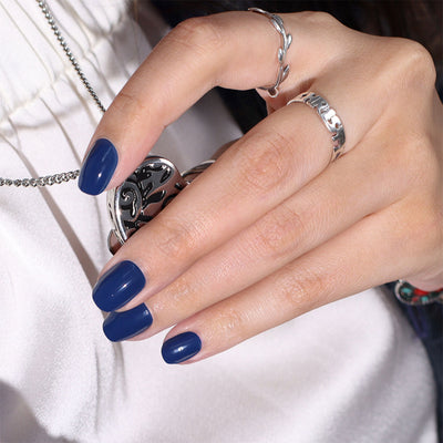 A hand with LONDONTOWN's "Under the Stars" blue-polished nails holds a silver locket necklace, complementing favorite silver rings on the ring and middle fingers. The blurred background adds mystery to this elegant look.