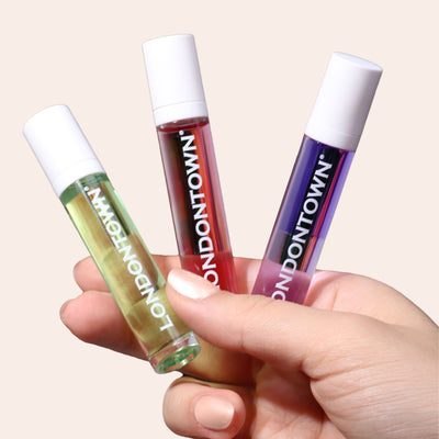 A hand holding a 3-piece Day to Night Cuticle Oil Set by LONDONTOWN, featuring glass bottles with white caps and filled with green, pink, and purple oils.