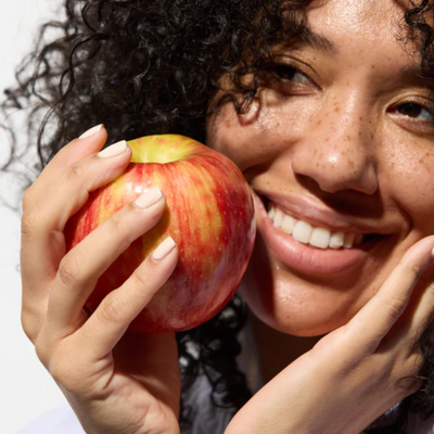 A person with curly hair smiles while holding LONDONTOWN Strengthening Nail Cream close to their face, sunlight casting soft shadows and highlighting the product's fresh features.