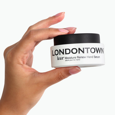 A hand holding a small white jar with a black lid labeled "Moisture Renew Hand Serum" by LONDONTOWN—a restorative solution for even the driest hands.