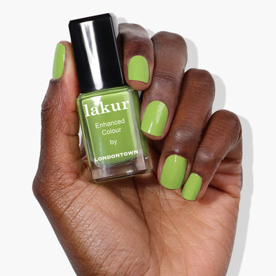 A hand with parakeet-green painted nails holds a bottle of LONDONTOWN Lakur Enhanced Colour nail polish in the shade Tipsy Mermaid.