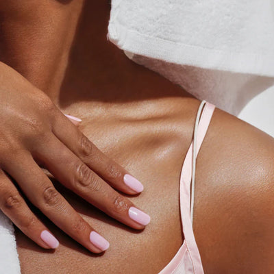 A person wearing LONDONTOWN's Petals & Pearls Set in light pink nail polish touches their shoulder, wrapped in a white towel and pink strap garment, showcasing everyday beauty.