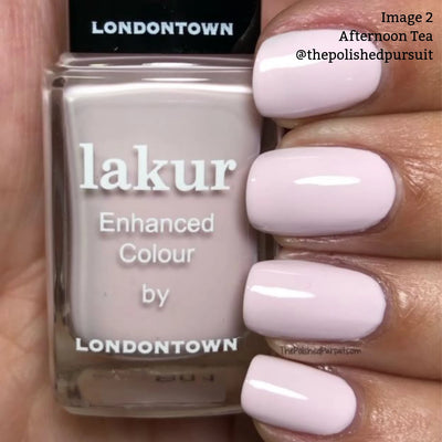Close-up of a hand with light pink crème nail polish in the shade “Afternoon Tea (Professional)” by LONDONTOWN. The bottle is labeled “lakur Enhanced Colour by LONDONTOWN” and is held against the painted nails, evoking a warm and cozy feeling reminiscent of a relaxing cup of tea.