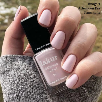 Close-up of a hand with light pink crème painted nails, holding a bottle of LONDONTOWN Afternoon Tea (Professional) nail polish. The hand, partially covered by a warm and cozy gray sweater, evokes the comforting feeling of enjoying a cup of tea.