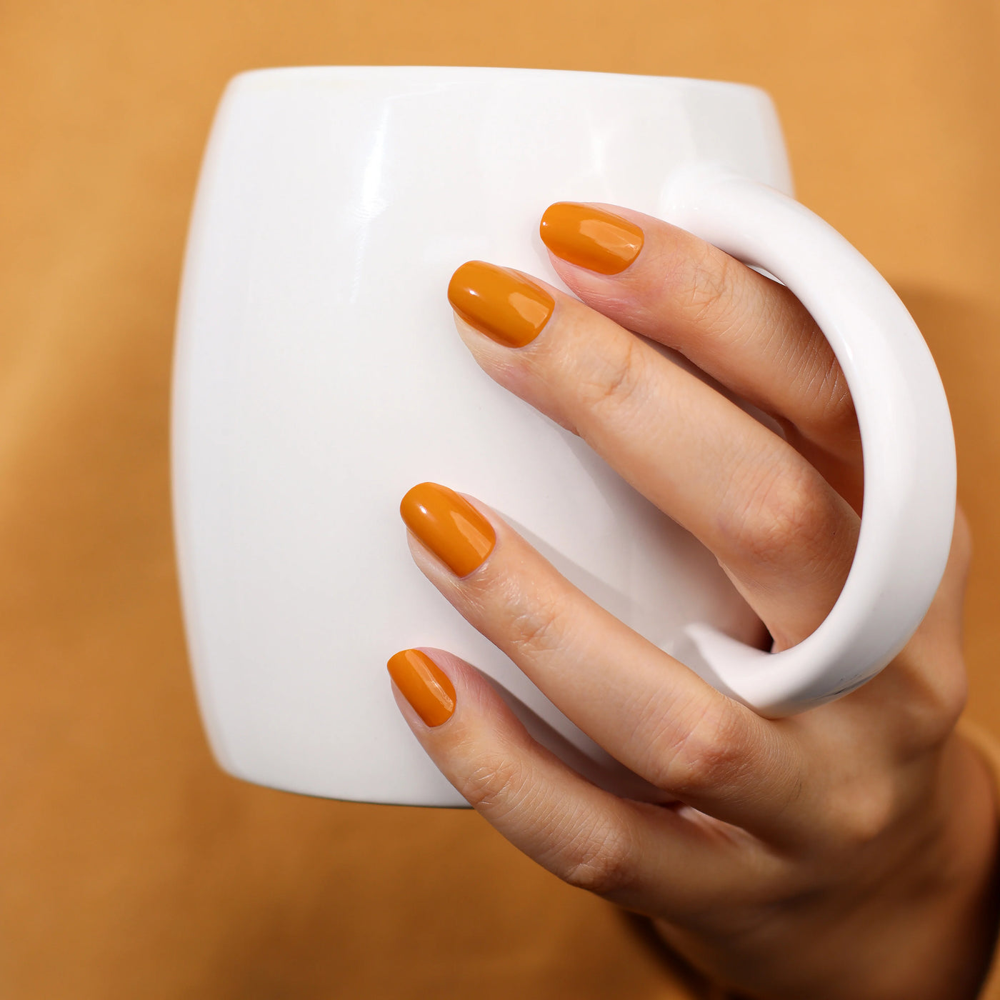 A hand adorned with nails painted in LONDONTOWN's Bee's Knees (Professional) nail polish holds a plain white mug against a robust, goldenrod background.