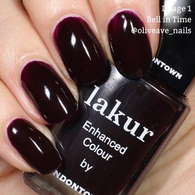 Close-up of a hand with fingernails painted deep purple, holding a bottle of nail polish labeled "Bell in Time (Professional)" by LONDONTOWN, known for its breathable formula and enriched with Florium Complex.