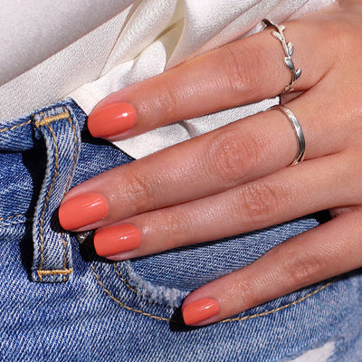 A hand with neatly manicured nails in LONDONTOWN's Pressed Petals, wearing two silver rings, rests on the waistband of blue denim jeans and a white blouse.