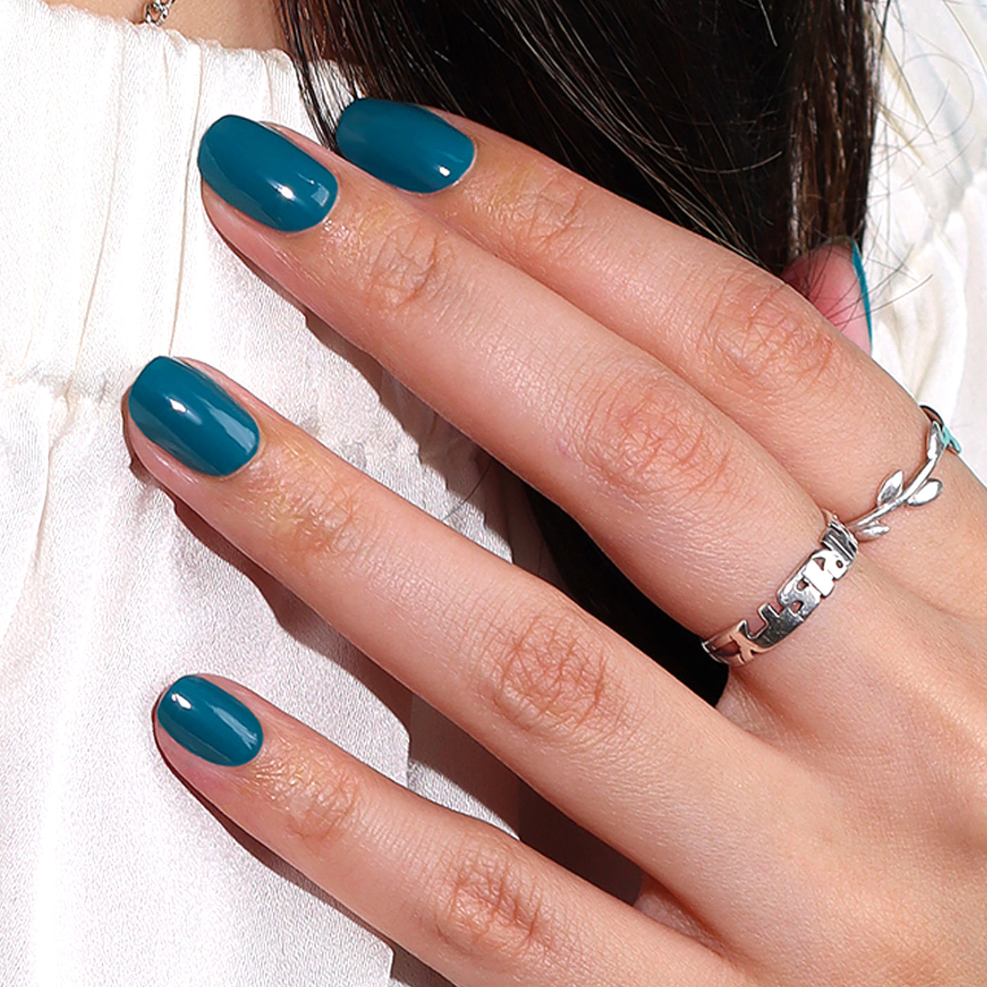 A hand with LONDONTOWN's "Magpie" peacock teal manicured nails and a silver ring rests against a white surface, making a sophisticated statement.