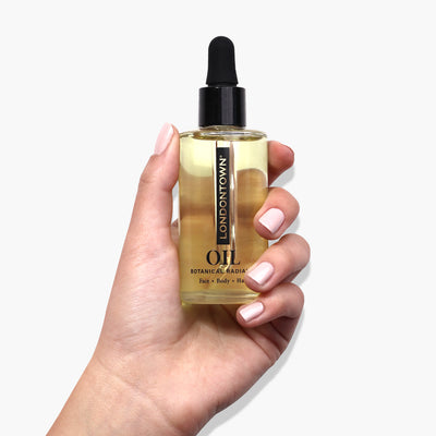 A hand holding a bottle of LONDONTOWN's Botanical Radiance Oil with a black dropper top against a white background, showcasing this multi-purpose oil.