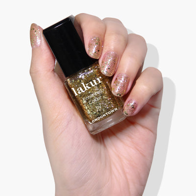 A hand with glittery pink nail polish holds a bottle of Britannia by LONDONTOWN, featuring a vegan, cruelty-free gold glitter polish with a transparent base.