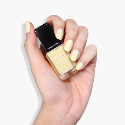 A hand with neatly manicured nails painted in a pastel yellow creme shade holds a bottle of "Buttercup" vegan, cruelty-free nail polish by LONDONTOWN.