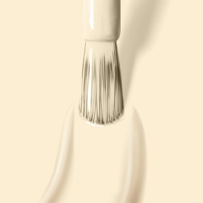 A close-up of a paintbrush applying creamy beige Buttercup by LONDONTOWN nail polish with bristles partially covered in the paint against a matching beige background evokes the smooth application of this 16+ Free, vegan, cruelty-free nail polish.