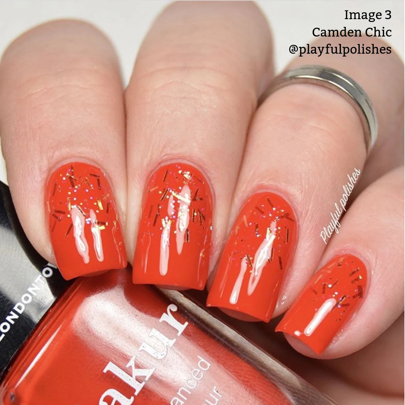 Close-up of a hand with glossy red vegan cruelty-free nail polish featuring glittery accents. The nail polish bottle reads "manakur" with the text "Image 3, Camden Chic (Professional) by LONDONTOWN @playfulpolishes" in the top right corner.