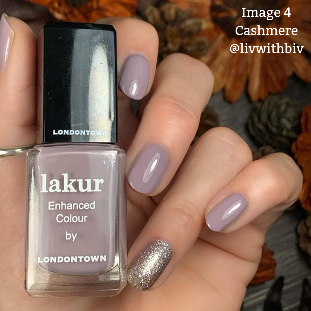 A hand with purple nail polish and a sparkling accent nail holding a bottle of LONDONTOWN Lakur Enhanced Colour, formaldehyde-free nail polish in the shade "Cashmere." The background features autumn leaves.