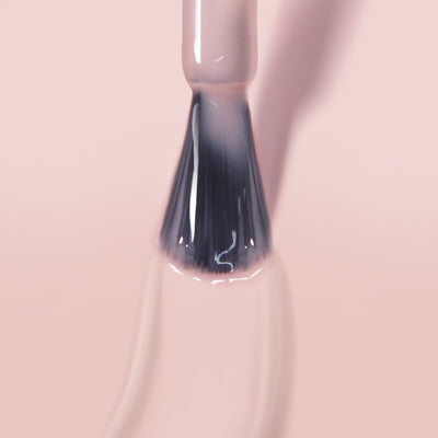 Close-up of a brush applying LONDONTOWN's Crowning Crumpet sheer nude nail polish on a light pink surface. The brush bristles are coated with the glossy gel manicure polish, leaving a shiny streak behind that benefits from color lock technology.