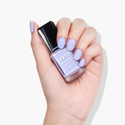 A hand holding a bottle of Lakur Enhanced Colour by LONDONTOWN in the shade "Dainty Daze," with nails painted in a matching periwinkle blue shade. This 16+ Free, vegan, cruelty-free formula ensures your manicure is both stunning and ethically responsible.