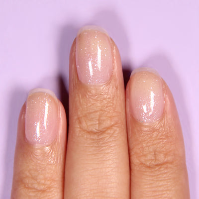 A close-up of fingers adorned with LONDONTOWN's Diamond Shine Top Coat, showcasing natural, glossy nails with a delicate diamond-dusted glittery finish against a light purple background.