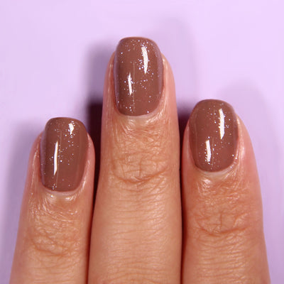 A close-up of three fingers with short, glossy, brown nail polish enhanced with LONDONTOWN's Diamond Shine Top Coat (Professional), featuring a diamond-dusted subtle sparkle effect. The background is a light purple color.