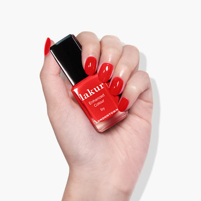 A hand with manicured red nails holds a bottle of LONDONTOWN's "Double the Deck" nail polish, showcasing its vibrant hue.