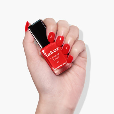 A hand with neatly manicured red nails holds a bottle of LONDONTOWN's "Double the Deck (Professional)," a true red crème vegan cruelty-free nail polish.