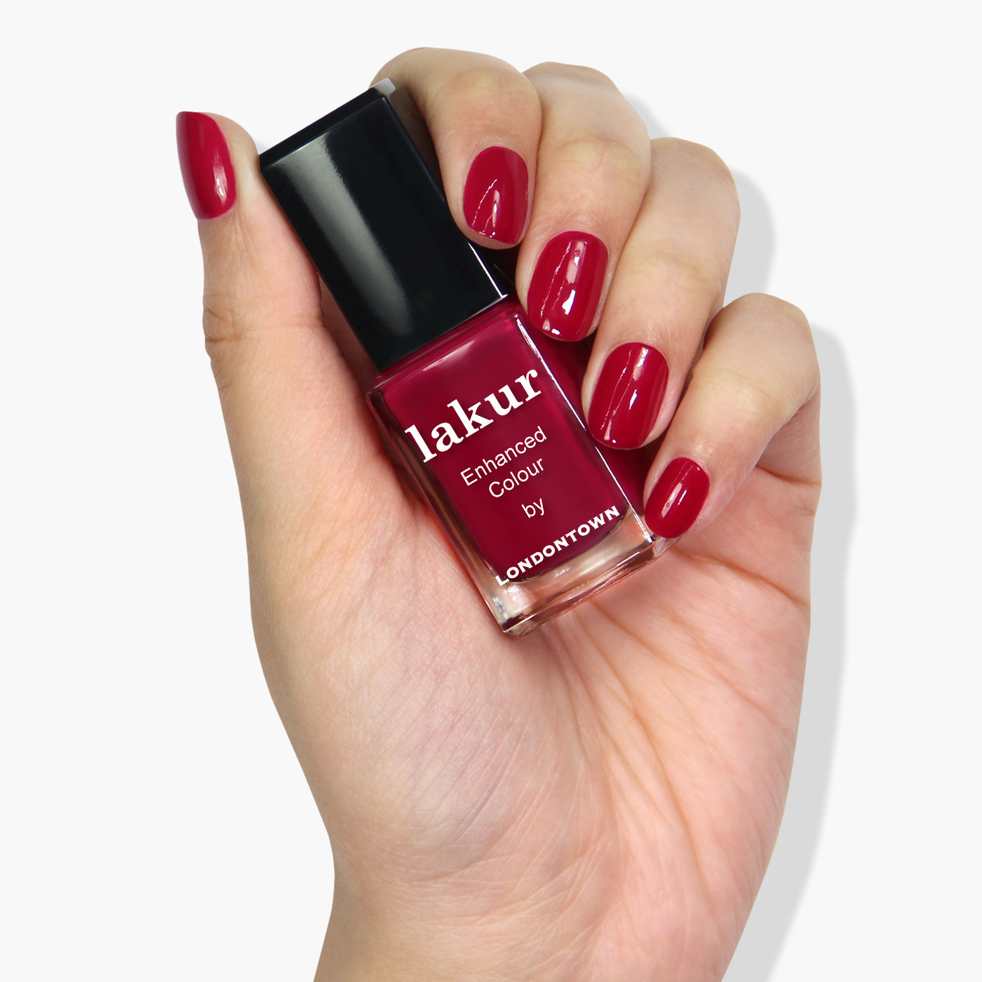 A hand with neatly manicured nails painted in deep red holds a bottle of LONDONTOWN's Dragon Fruit lakur enhanced colour nail polish, known for its cruelty-free and vegan formula.