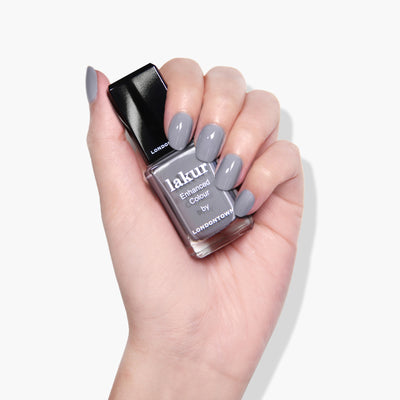 A hand with manicured nails painted in grey polish holds a bottle of LONDONTOWN's cruelty-free "Earl Grey" lakur Enhanced Colour nail polish.