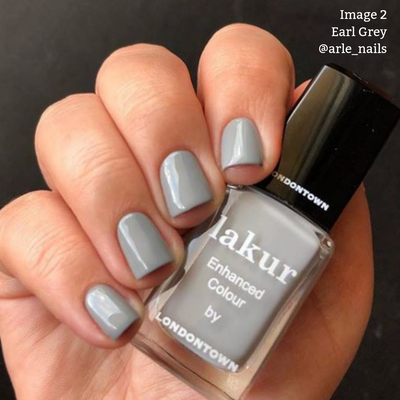 A hand with short, square-shaped nails painted in a pale grey shade holds a bottle of LONDONTOWN's Earl Grey Lakur Enhanced Colour nail polish. This cruelty-free and vegan polish is perfect for achieving an effortlessly chic look.