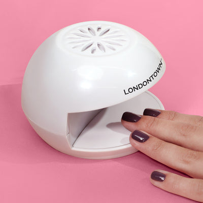 A hand with nails painted dark purple is placed under the "LONDONTOWN Flash Dry Nail Fan," a white semi-circular nail drying device, on a pink background—perfect for achieving salon-quality manis at home.