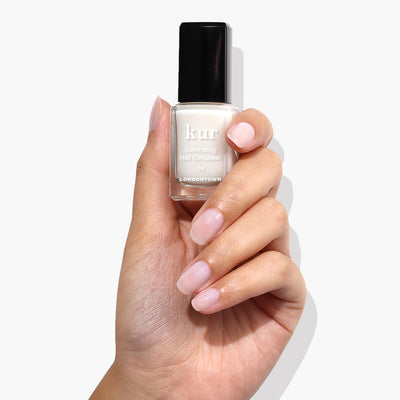 A hand holding a bottle of LONDONTOWN Illuminating Nail Concealer against a white background, showcasing polished nails with the perfect brightener for dull nails.