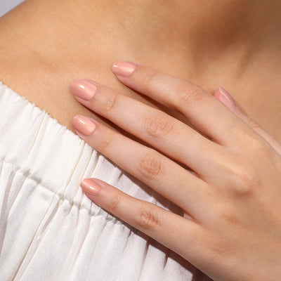 Close-up of a woman's hand with neatly manicured nails in LONDONTOWN's Amber Lights (Professional) shade resting on her shoulder, against a background of a white, off-the-shoulder top, reminiscent of a serene sunset.